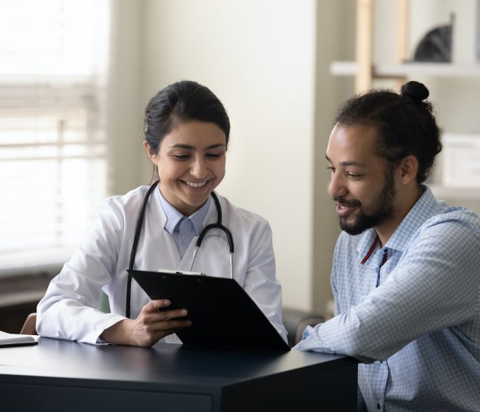 Happy young indian female gp doctor showing test result on clipboard to interested millennial african ethnicity male patient, discussing healthcare medical insurance or illness treatment at clinic.