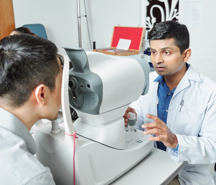 Indian serious doctor in white coat using medical equipment to examine his patient's eyesight at the hospital